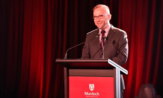 Adam Bandt in brown suit giving a speech on a Murdoch University podium