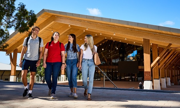 Four students walking toward the camera, talking and smiling, with large timber building in the background