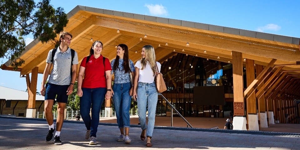 Four students walking toward the camera, talking and smiling, with large timber building in the background