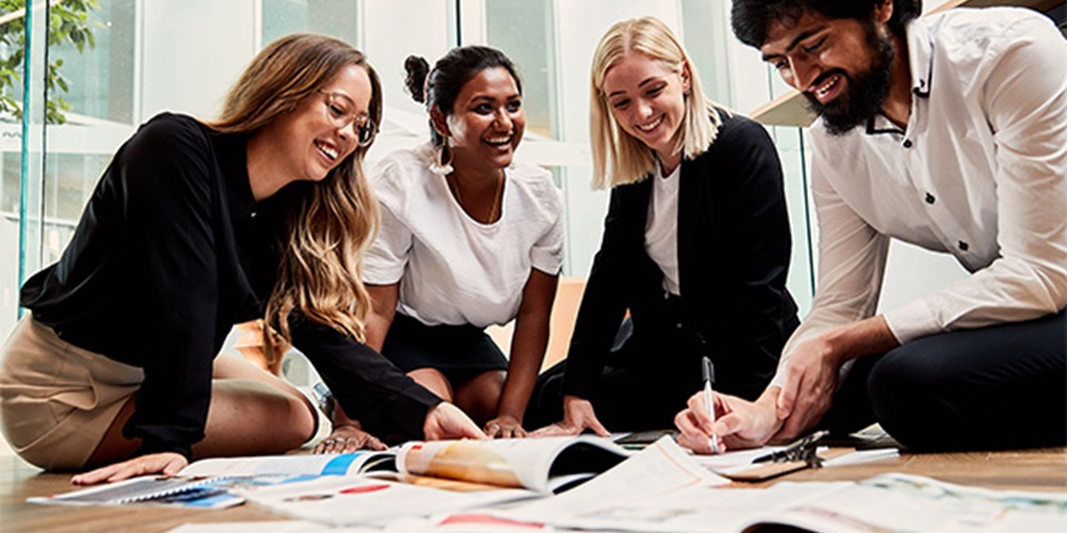 Four students sitting on the floor, smiling over an assortment of documents.