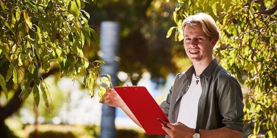 Student stands in bushland inspecting a bush with a clipboard