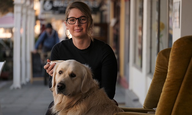 Ellie sits on a yellow couch with her golden retriever, both look at camera