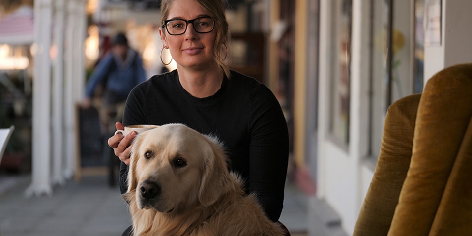 Ellie sits on a yellow couch with her golden retriever, both look at camera