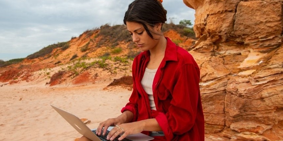 Student in red shirt sits on a red rock and studies laptop