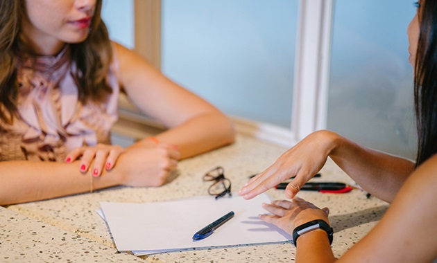 Two women sit at a table with a pen and paper between them