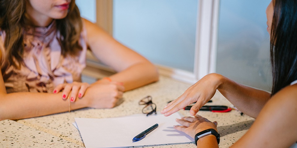 Two women sit at a table with a pen and paper between them