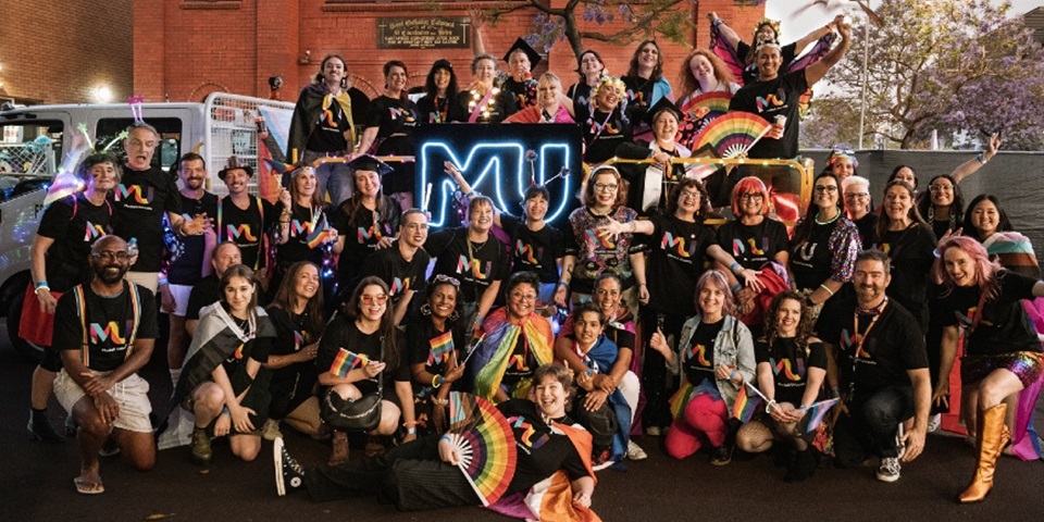 Large group of Murdoch University staff in black shirts with rainbow MU on front smiling at camera