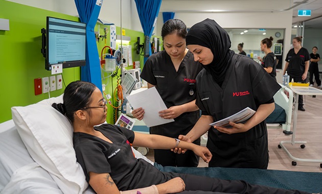 Two female students take the pulse of a third student who is lying on a bed in the SimLab.