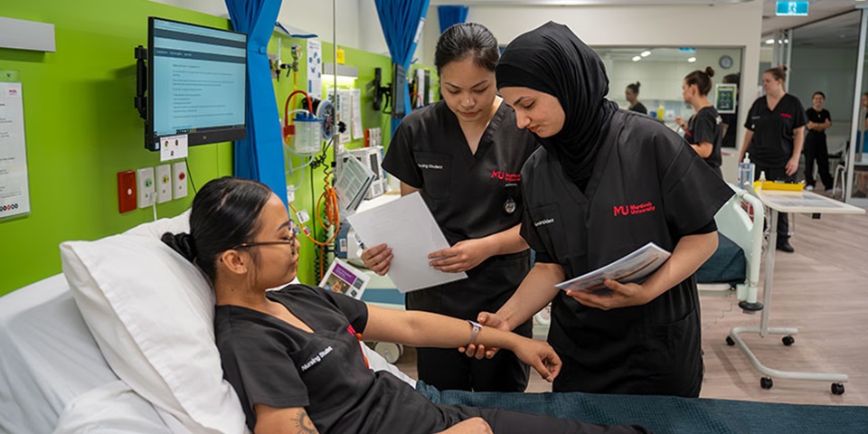 Two female students take the pulse of a third student who is lying on a bed in the SimLab.