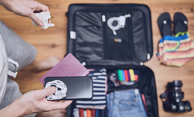 Man standing over suitcase filled with clothes holding a phone, passport, and adaptor