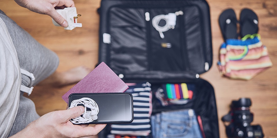 Man standing over suitcase filled with clothes holding a phone, passport, and adaptor