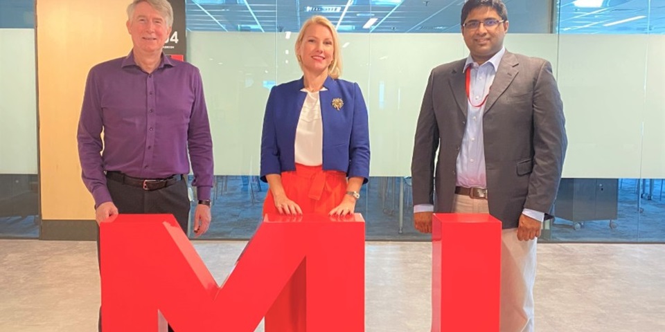 Dr James Trotter, Monika Fourneaux and Joseph John Nalloor standing behind a large MU sign