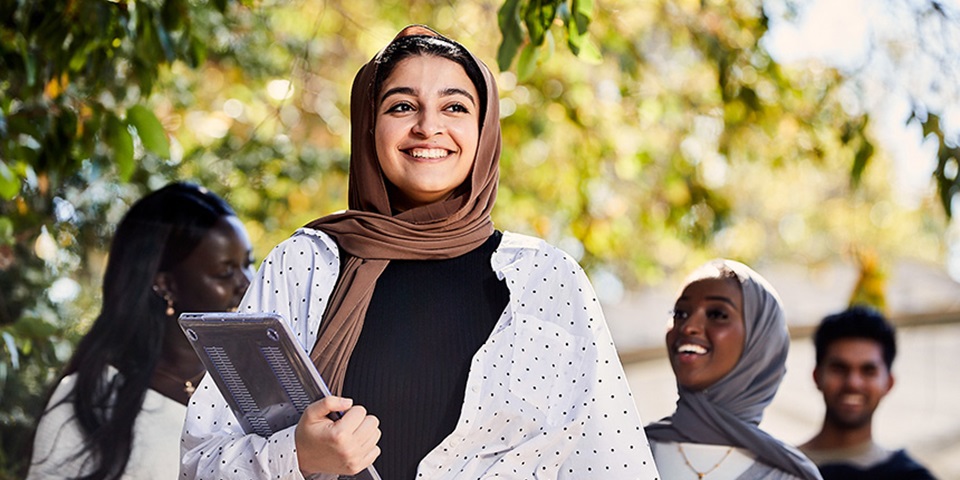 Female student wearing hijab smiling and holding laptop.